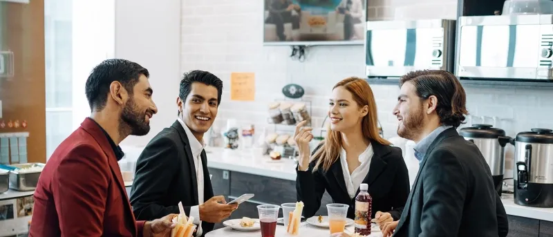 Four diverse business professionals—two men in suits, one man in a blazer, and one woman—sit at a table in a bright cafeteria or break room, talking and smiling during a lunch break.